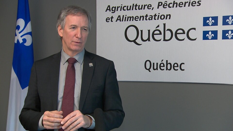 André Lamontagne devant une affiche du ministère et un drapeau du Québec lors d'une entrevue.