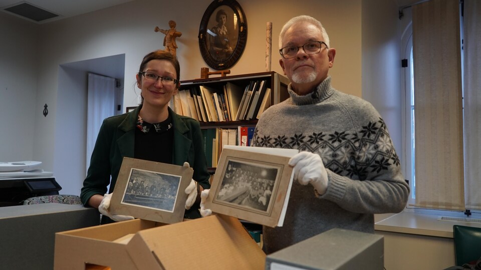 Une femme et un homme tiennent dans leurs mains des photos historiques de la Beauce. Des boîtes ouvertes se trouvent devant eux.