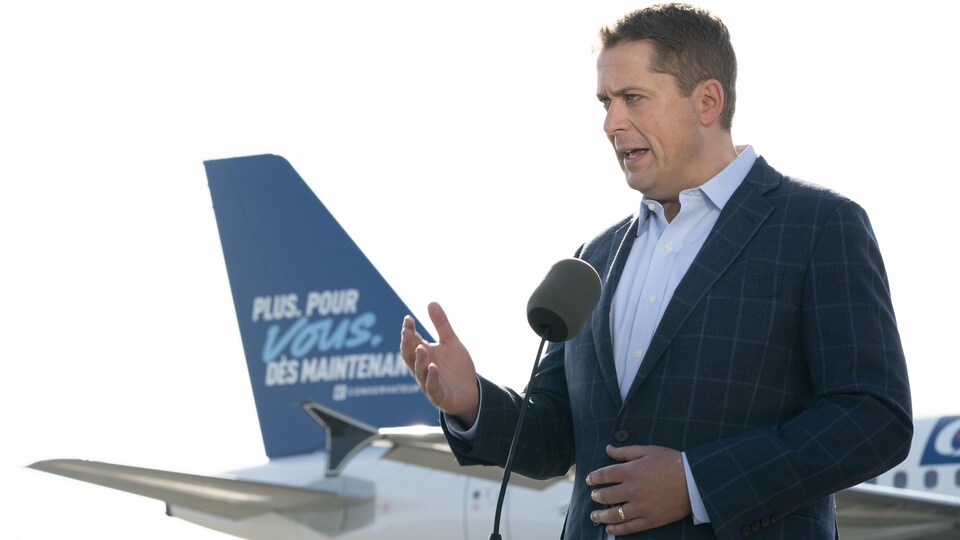 Andrew Scheer, devant la queue d'un avion sur laquelle on peut lire le slogan du Parti conservateur.