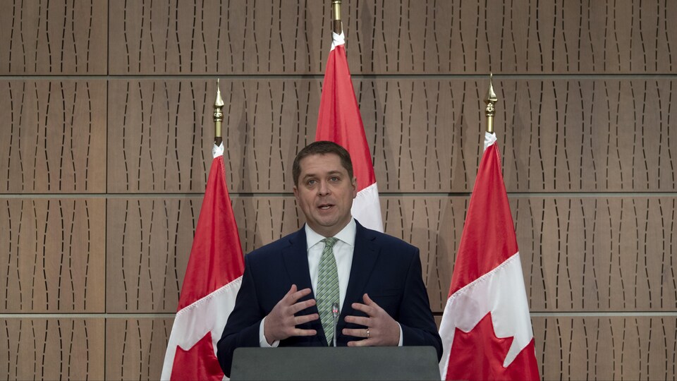 Andrew Scheer parle devant trois drapeaux du Canada.