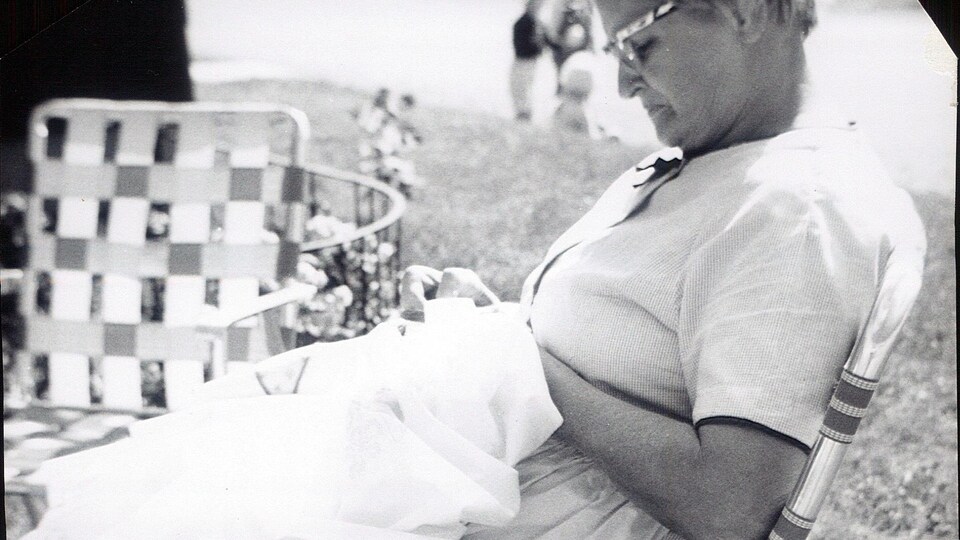 Une photo ancienne de la grand-mère de l'artiste visuelle fransaskoise Anne Brochu Lambert. Elle fait de la couture assise dans une chaise à l'extérieur.