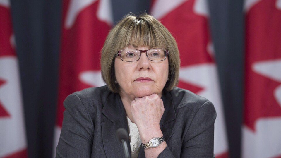 Une femme assise, le menton appuyée sur son poing fermé, regarde au loin. On voit des drapeaux du Canada derrière elle.