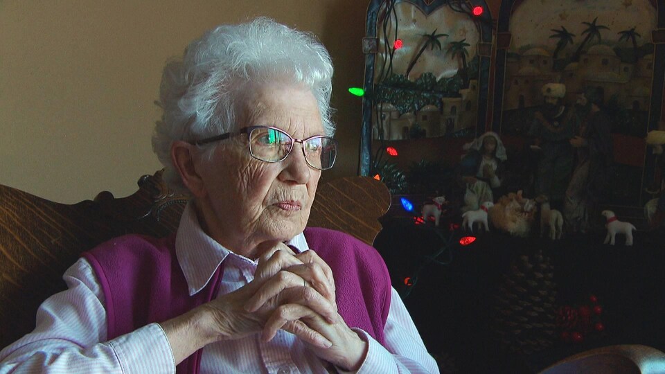 Une femme de 90 ans les mains jointes, assises près d'une crèche de Noël entourée de lumières.
