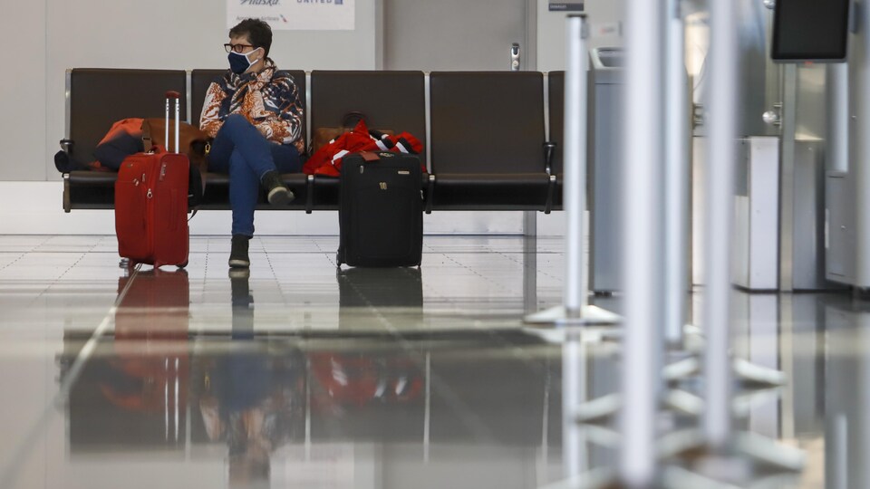 Une dame assise à l'aéroport avec ses valises. Elle porte un masque.