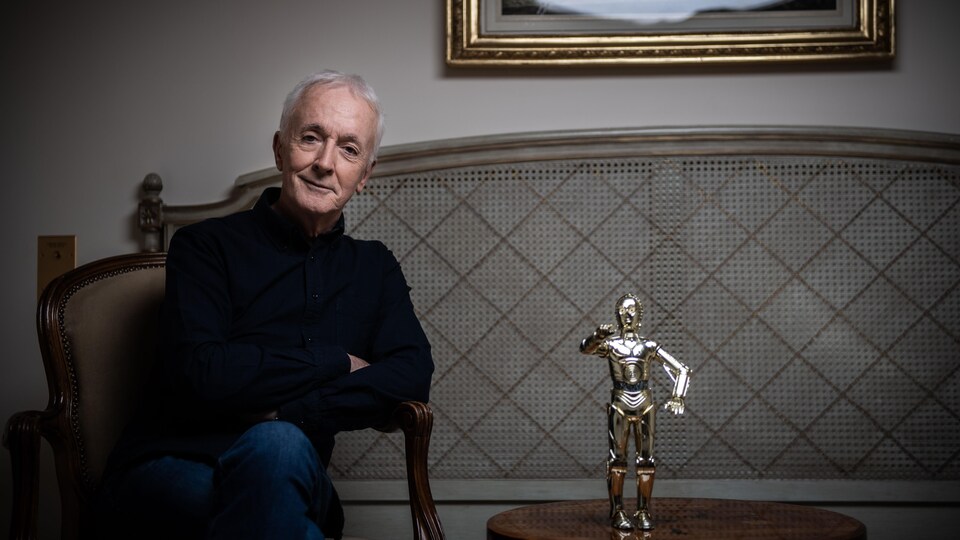L'acteur est assis sur un fauteuil, devant une table où trône une figurine de C-3PO, le personnage qu'il incarne dans les films.