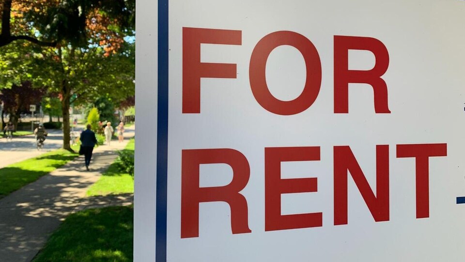 Un passant marche sur le trottoir et croise une affiche sur laquelle les mots « For Rent » sont écrits en rouge.