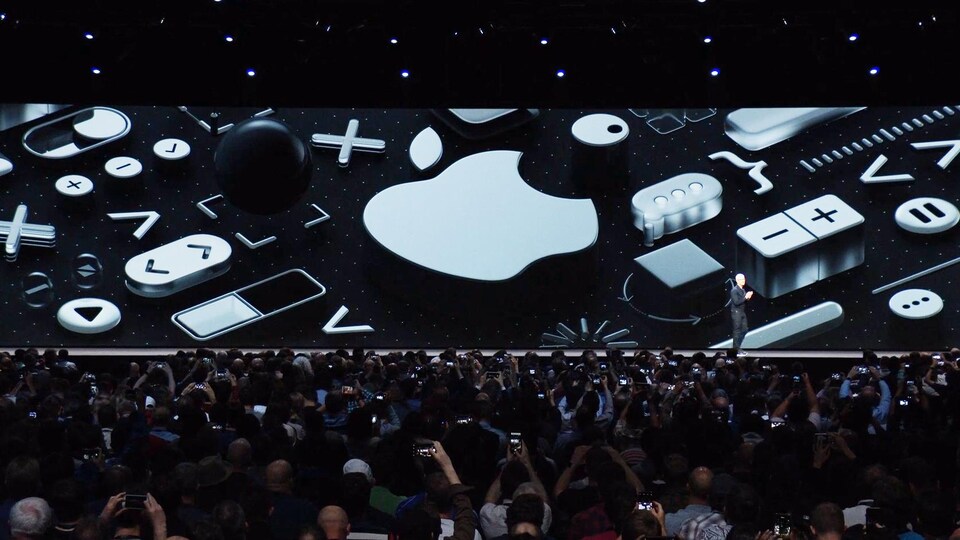 Une capture d'écran de la présentation des nouveaux logiciels d'Apple montrant une vue d'ensemble de la salle, avec un énorme écran sur la scène, derrière Tim Cook.