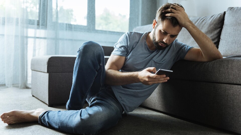 Un homme triste consulte son téléphone. Il est assis sur le tapis de son salon consulte son cellulaire.