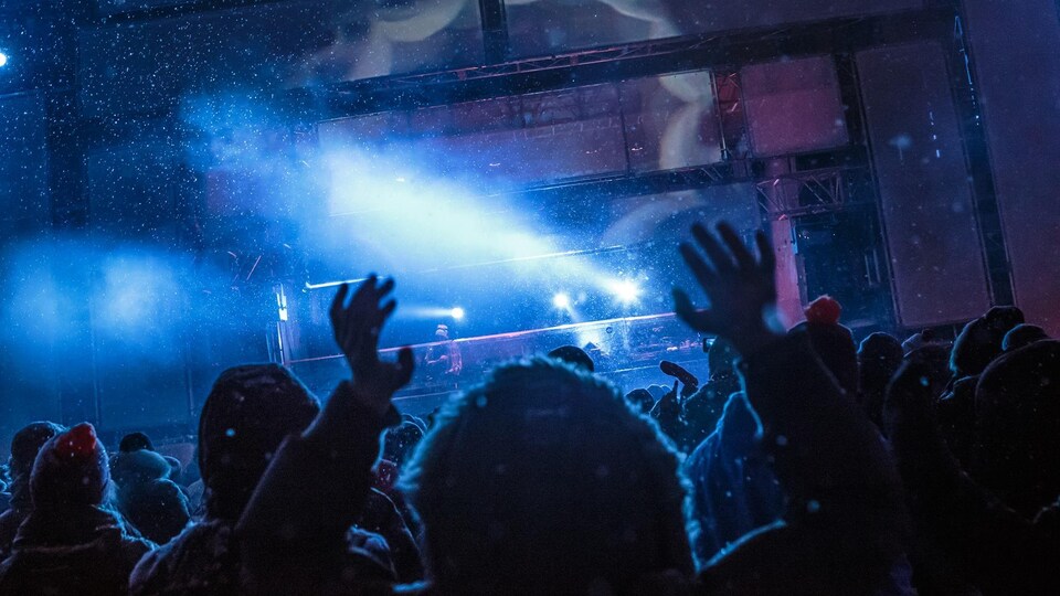 Des gens de dos lèvent les bras en regardant un artiste performer sur une scène extérieure, pendant une nuit enneigée. 