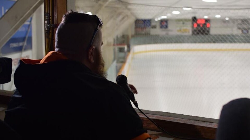 Un homme qu'on voit de dos dans une cabine regarde la patinoire d'un aréna. 