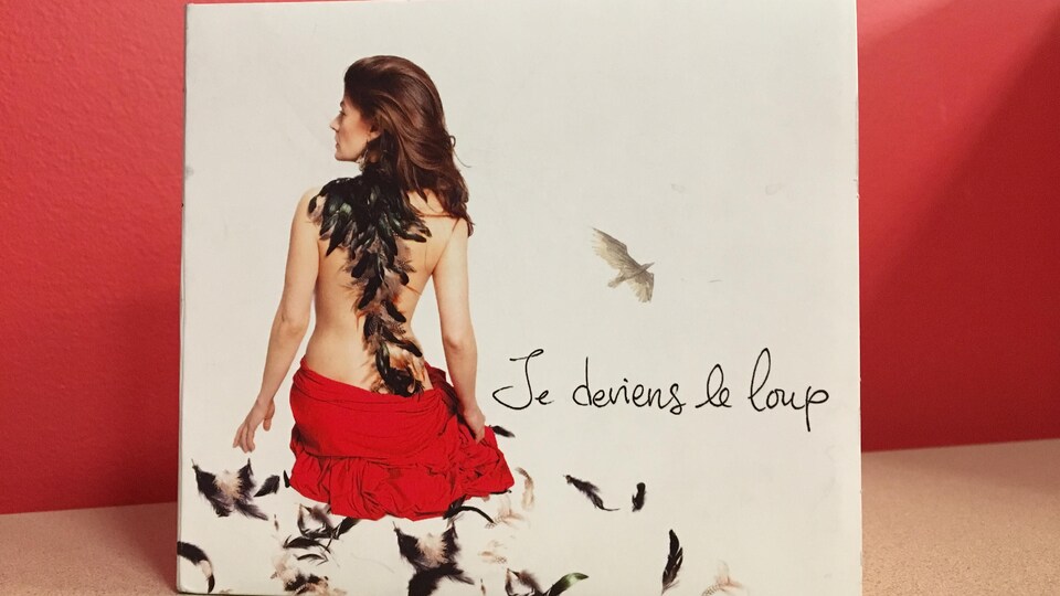 Un album d'un disque compact est devant un mur rouge. Sur la pochette, une femme a le dos nu et des plumes volent au vent.