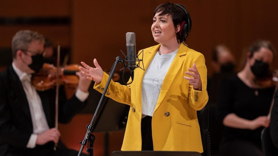 La femme chante sur scène derrière un micro. On aperçoit derrière elle des violonistes.