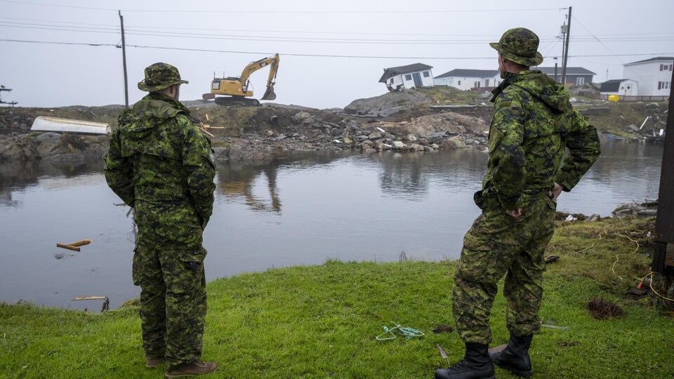 Deux militaires en uniforme au bord de l'eau observent une excavatrice et une maison qui menace de tomber, sur la rive opposée.