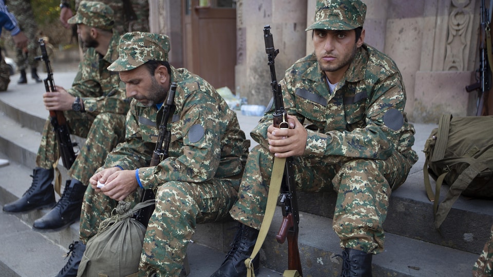 Des militaires attendent, assis sur des marches dehors en tenant leur fusil. 