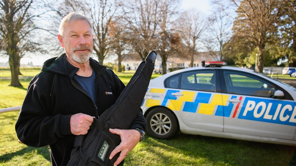 Un homme tient un sac de transport d'arme devant une voiture de police.