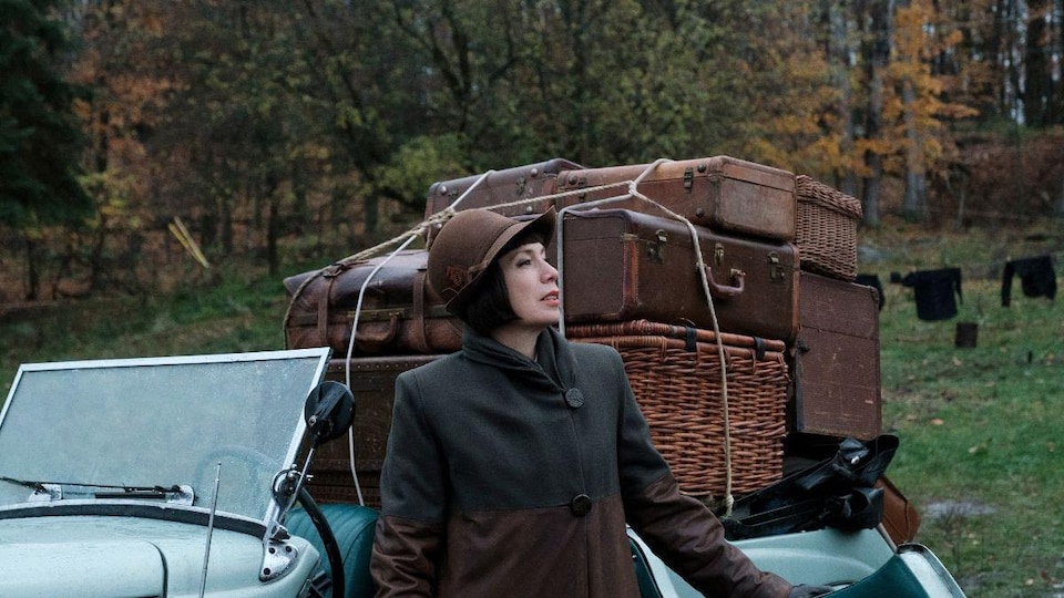 L'actrice, en vêtements d'époque, se tient devant une vieille voiture remplie de bagages. 