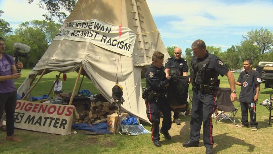 Trois policiers soulèvent du sol un manifestant, il y a des gens autour, ils sont tous devant le tipi installé au parc Wascana.