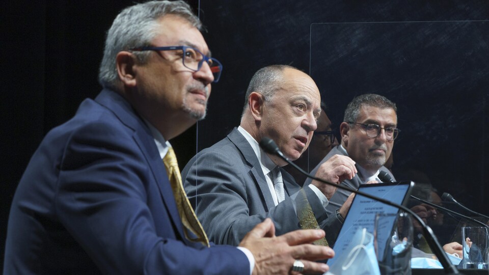 Trois hommes assis à une table en conférence de presse.