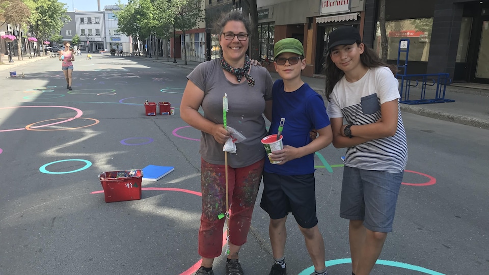 Une artiste et deux adolescents posent au centre de la rue où ils ont peint des motifs au sol.