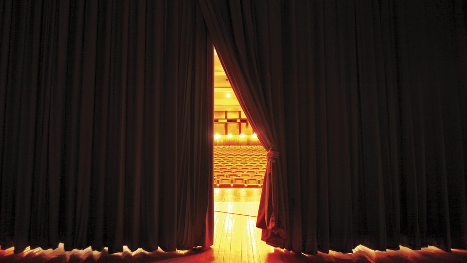 Un rideau de théâtre rouge, entrouvert montre la salle éclairée et des sièges vides.