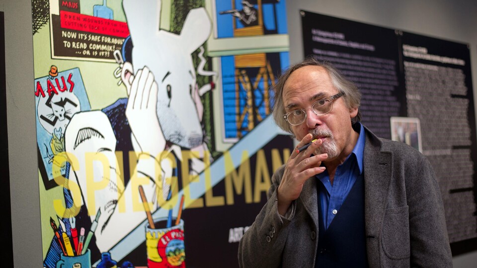 Un homme fume un petit cigare devant une planche de sa bande dessinée montrant une souris qui fume.