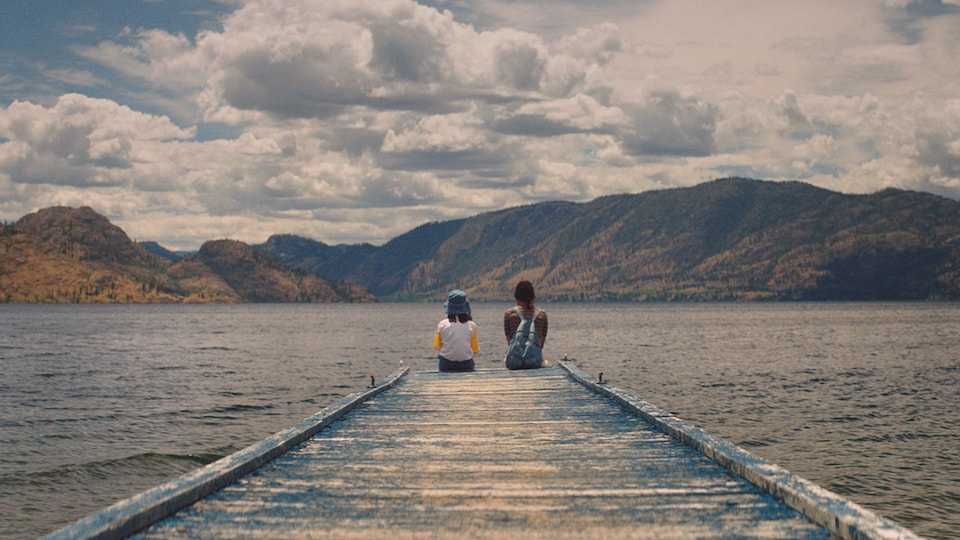 Two people are sitting on the edge of a dock and looking at the water and the mountains around them