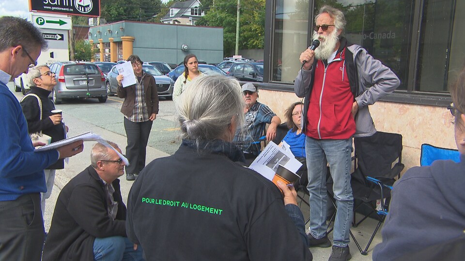 Un homme barbu s'adresse à quelques personnes avec un micro devant les bureaux de radio-canada.