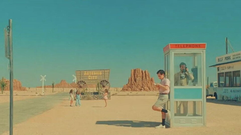 Un homme est adossé contre une cabine téléphonique située dans le désert, dans laquelle un homme parle au téléphone, alors que des enfants jouent au loin. 