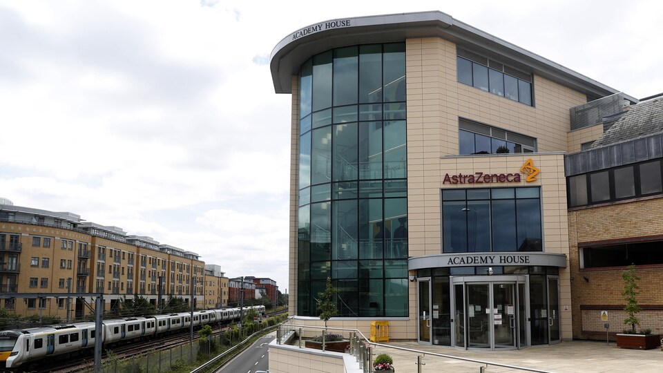 Un immeuble moderne avec le logo d'AstraZeneca en façade, près d'une voie où circule un train dans un quartier résidentiel.