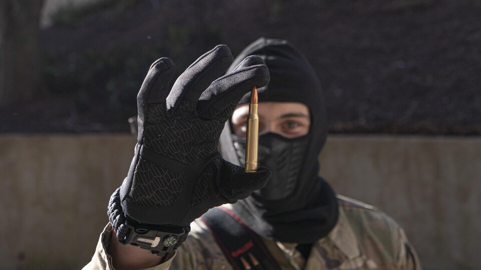 Un homme cagoulé, masqué, en habit de camouflage, tient une balle de fusil entre son pouce et son index.