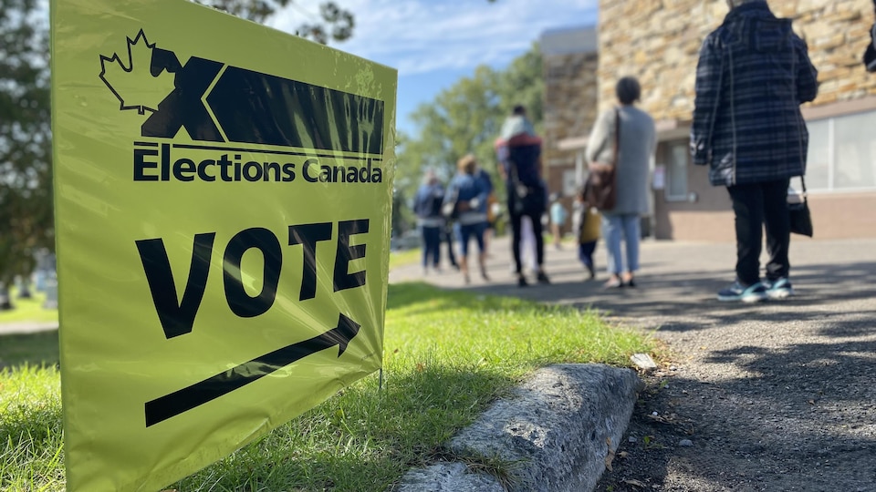 Une affiche indiquant la direction d'un bureau de vote en avant-plan et une file d'attente en arrière-plan.