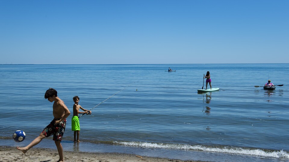 Un garçon joue au ballon sur la plage, un jeune garçon pêche et sur l'eau des gens s'amusent sur des planches. 