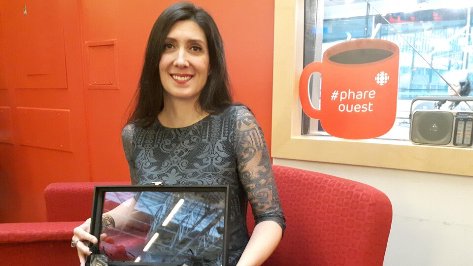 Une femme assise sur un fauteuil devant une fenêtre tient un coffret ouvert dans lequel reposent des bagues ornées de verre peints.