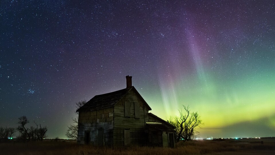 Une maison abandonnée se dresse devant un ciel étoilé et illuminé par des faisceaux lumineux multicolores.