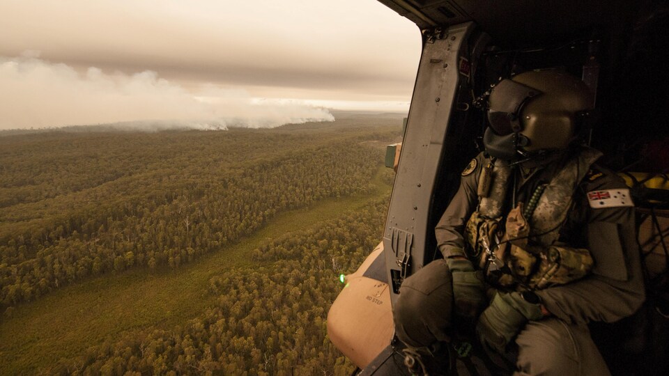 Un militaire portant un masque respiratoire est assis dans un hélicoptère et regarde la fumée qui plane au-dessus d'une forêt.