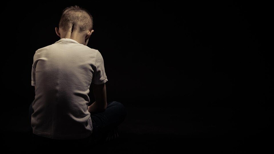 Dans un pièce noire, un enfant autiste de dos la tête baissée. 