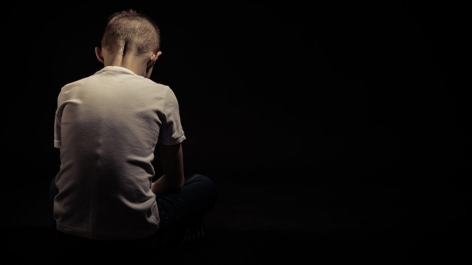 Dans un pièce noire, un enfant est vu de dos, la tête baissée.