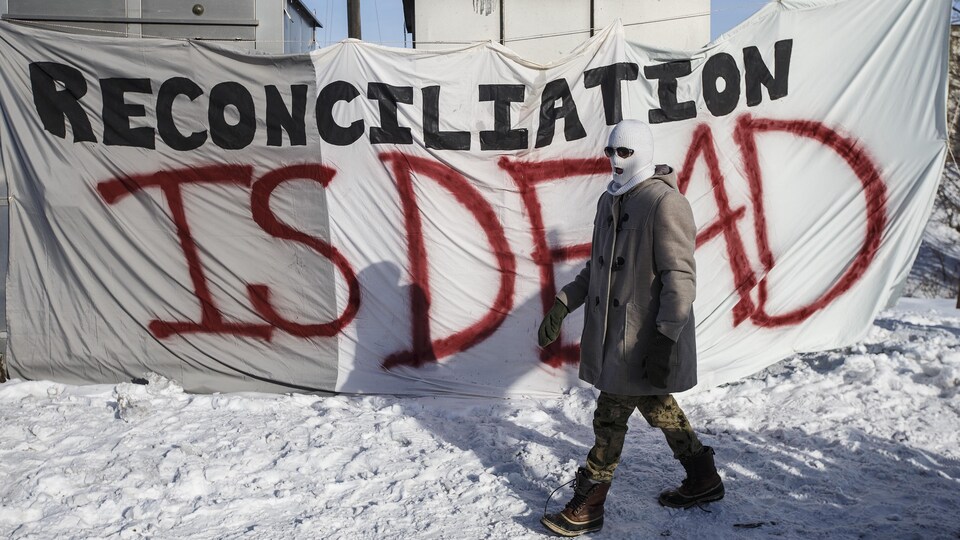 Une personne masquée devant une banderole sur laquelle est écrit : « Reconciliation is dead ».