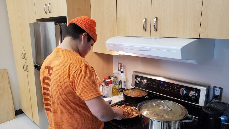 Un homme autochtone est dans une cuisine, on y voit deux chaudrons et une poêlonne sur un four avec de la nourriture dedans. 