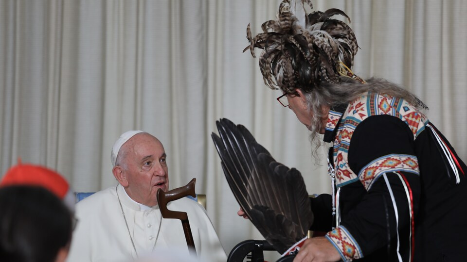 Un Autochtone portant une tenue traditionnelle munie de plumes s'adresse au pape qui est assis devant l'auditoire.