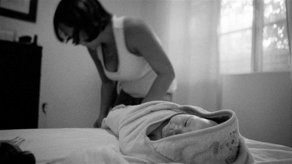 Une image en noir et blanc d'une femme en arrière-plan avec en avant-plan un bébé emmailloté sur un lit dans une chambre à coucher.