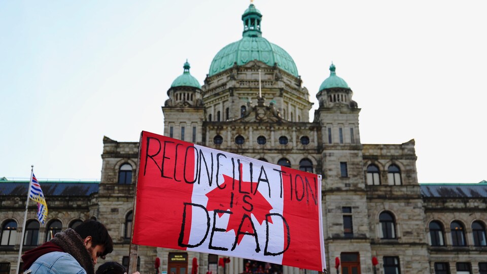 «La réconciliation est morte» lit-on sur un drapeau canadien, porté par un manifestant devant l'Assemblée législative de la C.-B.
