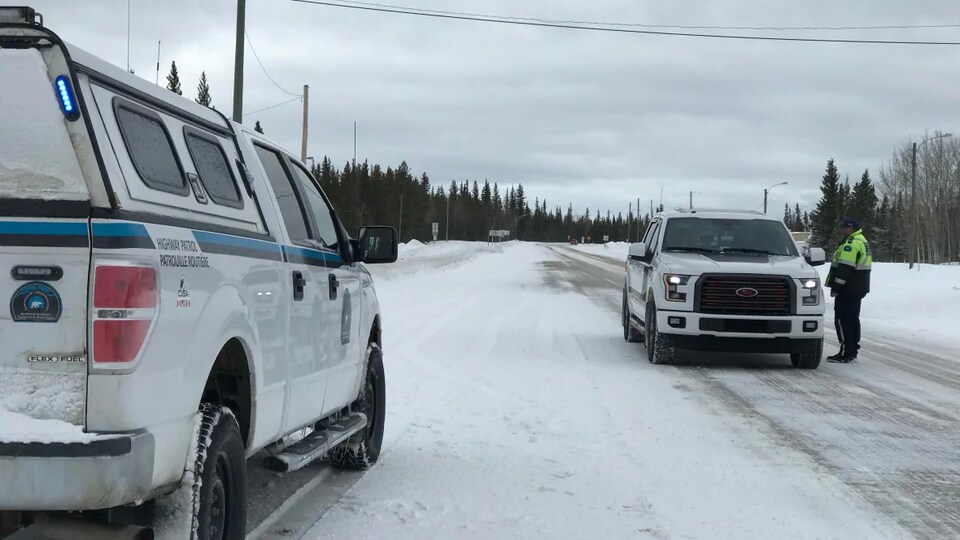 Un agent de police est debout près d'un véhicule arrêté sur le bord d'une route enneigée bordé de forêts.