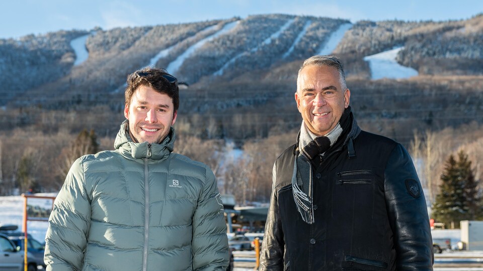 Alex Harvey et Mario Bédard posent devant la montagne du Mont-Sainte-Anne. On voit des pistes de ski au-dessus de leurs têtes. 