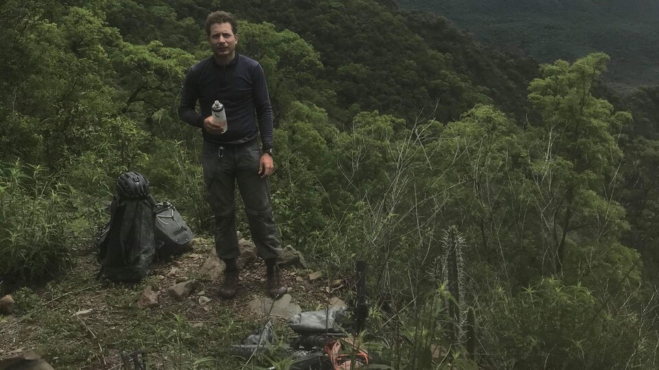 Frédéric Dion pose sur une falaise donnant sur une grande vallée, avec son sac à dos et son vélo à côté de lui.