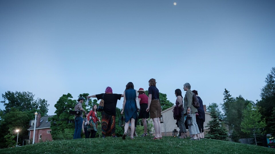 Un groupe formé d'une dizaine de personnes formé d'acteurs du spectacle et des membres du public sur une petite bute avec de la pelouse à la tombée de la nuit.