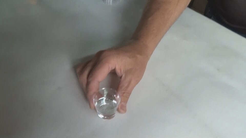 Une main dépose sur une table un petit verre contenant un liquide clair.