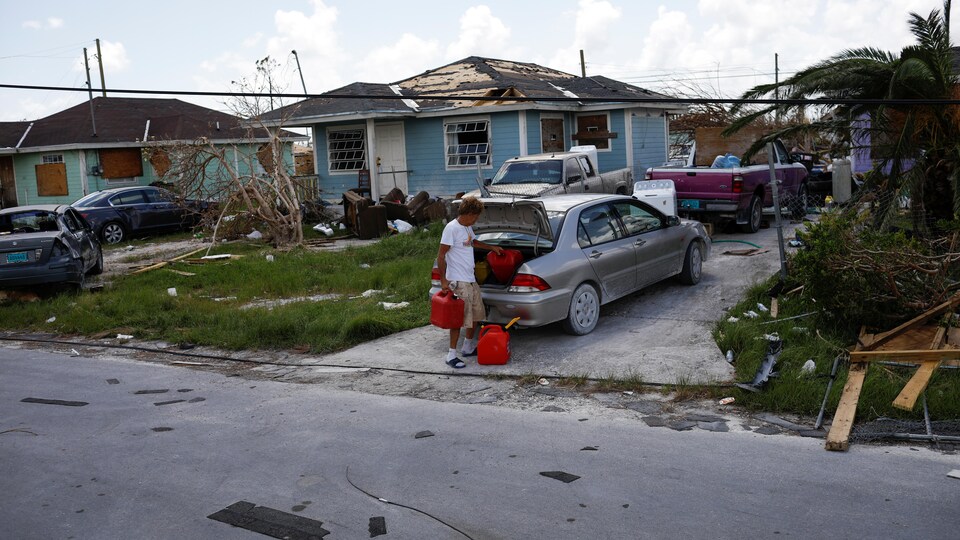 Un homme sort les bidons du coffre d'une voiture, dans un voisinage dont les maisons sont endommagées.