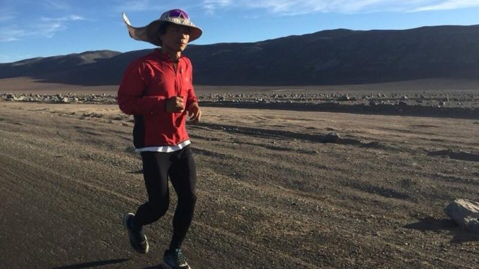 Bai Bin court dans un secteur désertique et caillouteux avec un chapeau sur la tête sous un ciel partiellement nuageux.