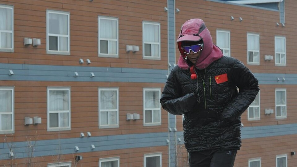Bai Bin court près d'un immeuble à Inuvik avec des lunettes de soleil et un manteau sur lequel il a collé un petit drapeau chinois.